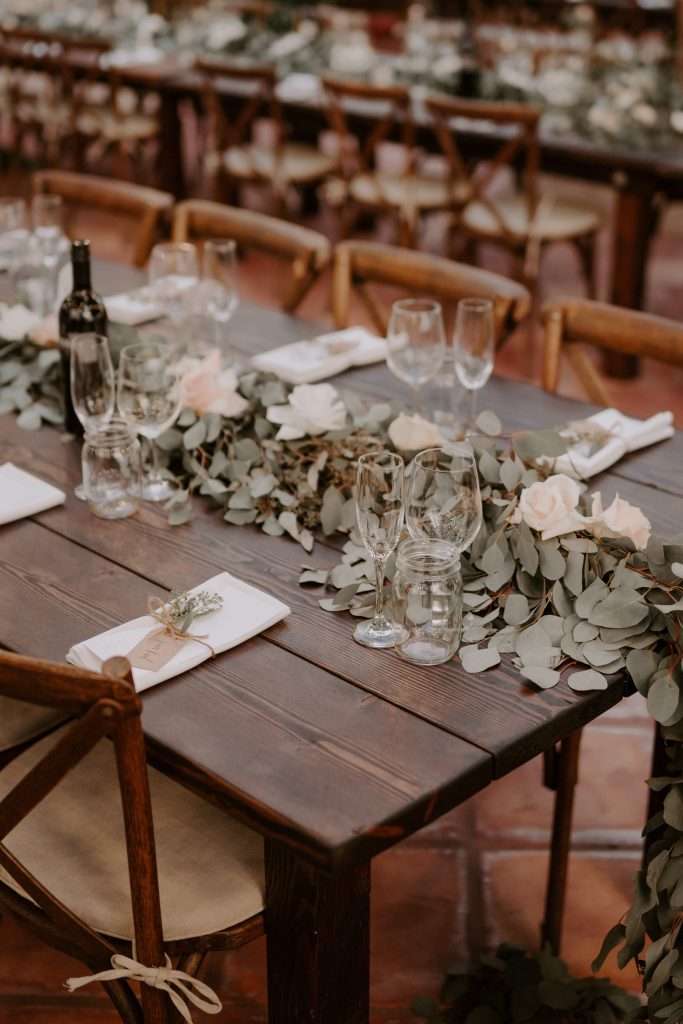 Dark wood farm table with a eucalyptus runner and cross-back chairs set for a wedding dinner